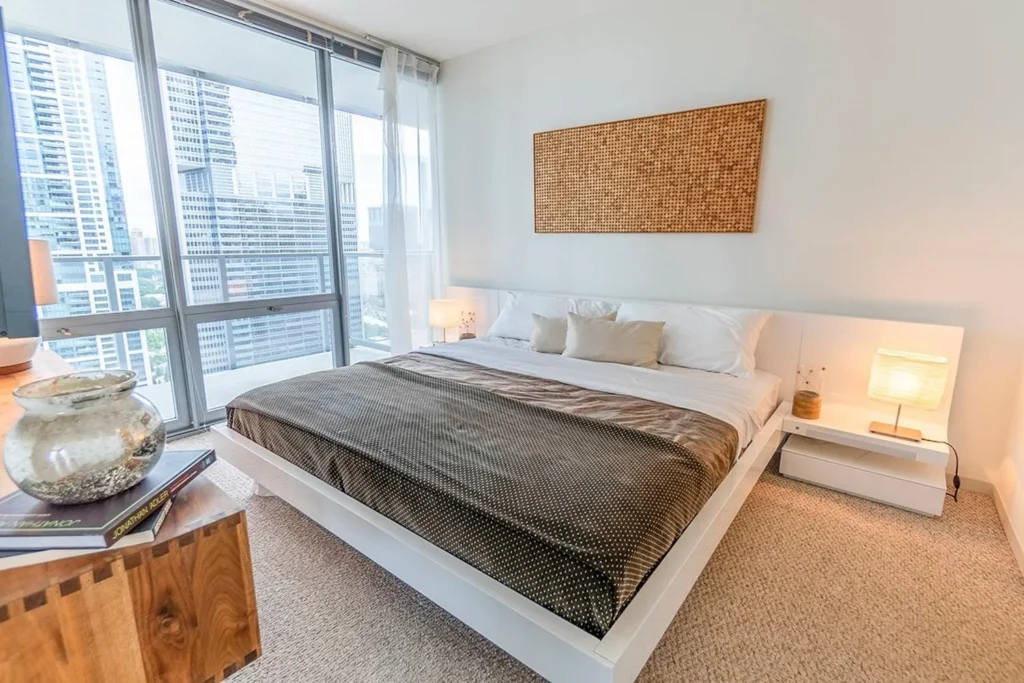 Modern bedroom with a large bed, two nightstands with lamps, a woven wall art above the bed, and floor-to-ceiling windows overlooking city buildings.