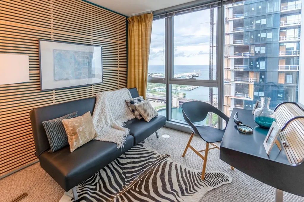 Modern living room with a black futon, desk, zebra rug, and large windows overlooking a cityscape and waterfront.