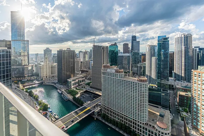 Aerial view of downtown Chicago with high-rise buildings, the Chicago River, and a bridge under a partly cloudy sky.