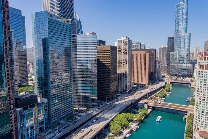 Aerial view of downtown Chicago skyscrapers along the Chicago River on a clear, sunny day, with bridges spanning the water and boats visible below.