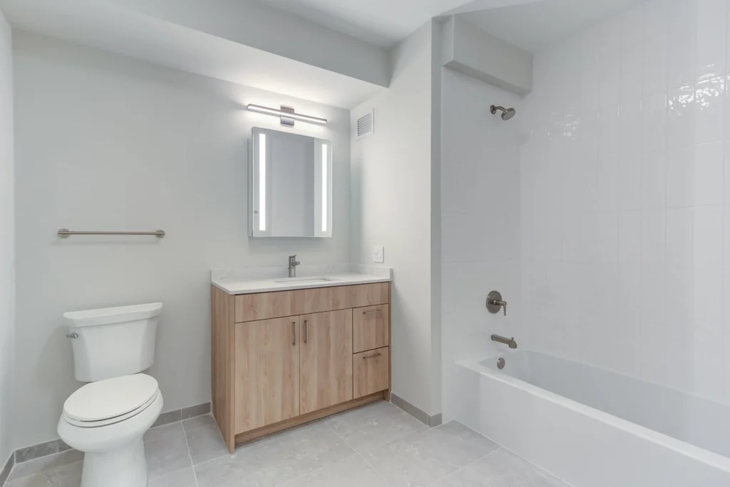 Modern bathroom with a toilet, wooden vanity with a sink and mirror, wall-mounted light, and a bathtub with a showerhead, all in neutral tones.