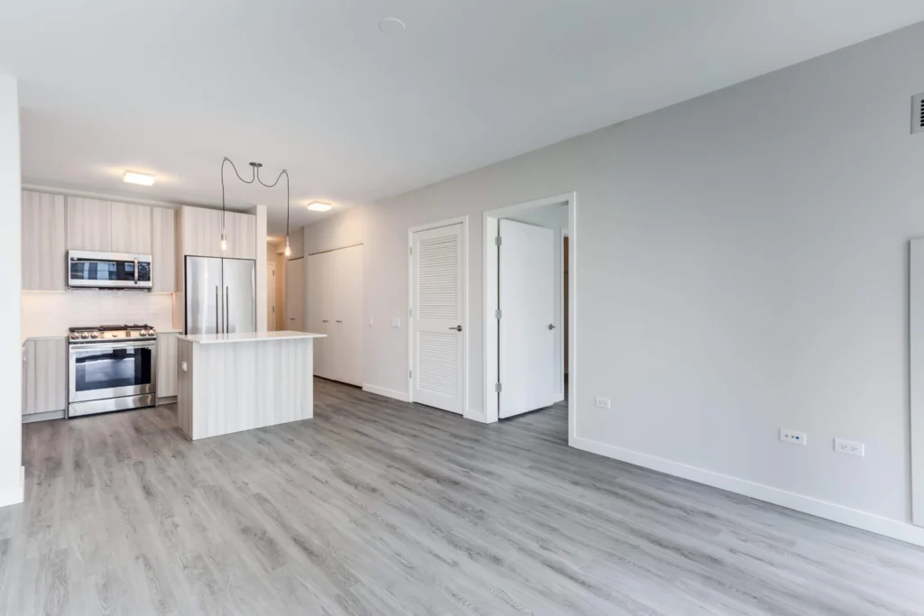 Modern kitchen and living area with light wood floors, stainless steel appliances, an island, and adjacent doorway leading to other rooms.