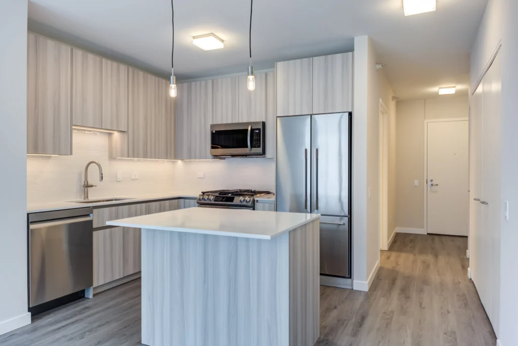Modern kitchen with light wood cabinets, stainless steel appliances, island with white countertop, pendant lights, and wood flooring. Entry hallway visible in the background.