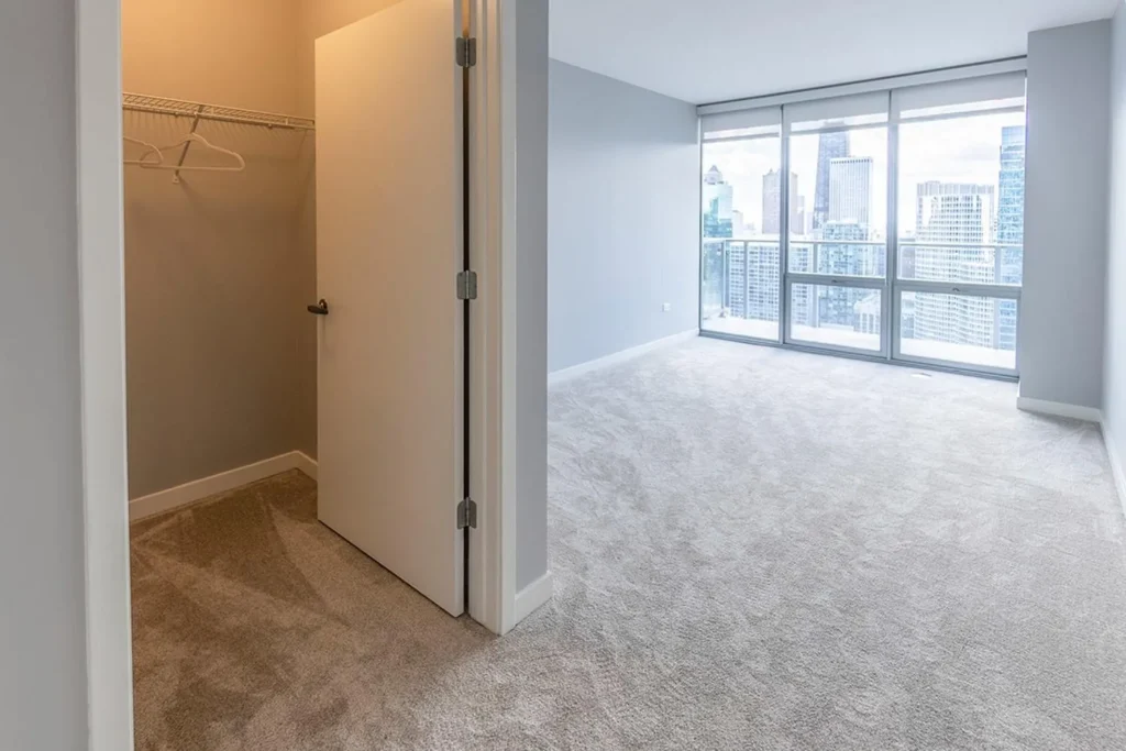 A carpeted bedroom with large windows and a city view, next to an open closet with a wire shelf and a white door.