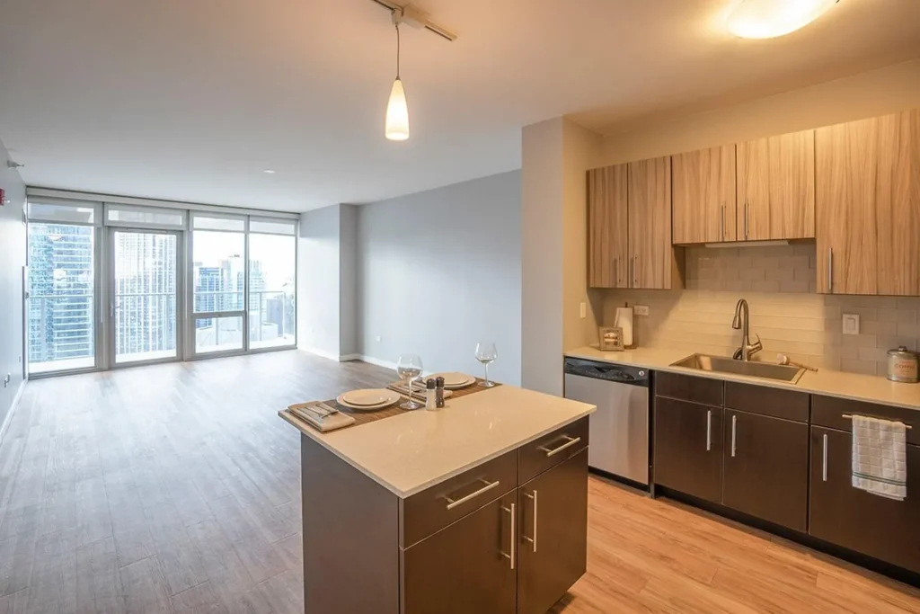 Modern apartment interior with open kitchen, island, and living area; large windows offer a city view, and the space features wood-style flooring and neutral-colored cabinets.