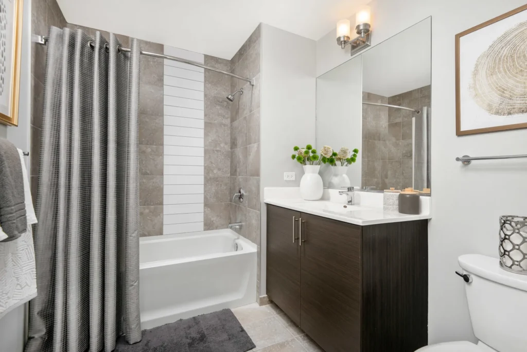 Modern bathroom with a tiled shower and tub, a dark wood vanity with a white sink, large mirror, toilet, wall art, and neutral decor accents.