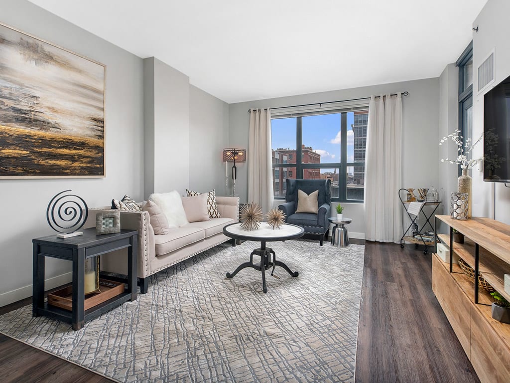 Modern living room with a beige sofa, abstract artwork, round coffee table, and large window with city view. Neutral tones and contemporary decor elements throughout the space.