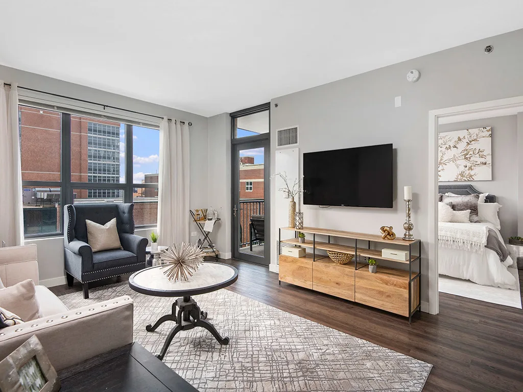 Modern living room with large windows, a wall-mounted TV, wooden console, neutral furniture, and a view into a bright bedroom.