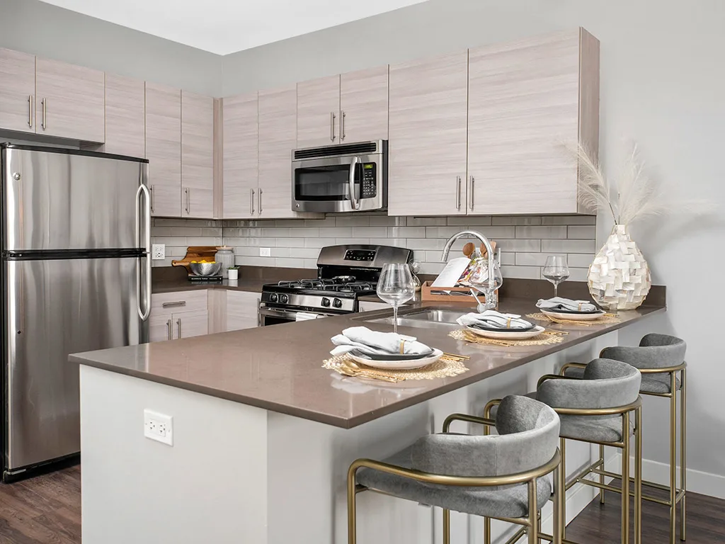 Modern kitchen with light wood cabinets, stainless steel appliances, a brown island countertop, three gray barstools, and neatly set place settings with glassware and decorative accents.