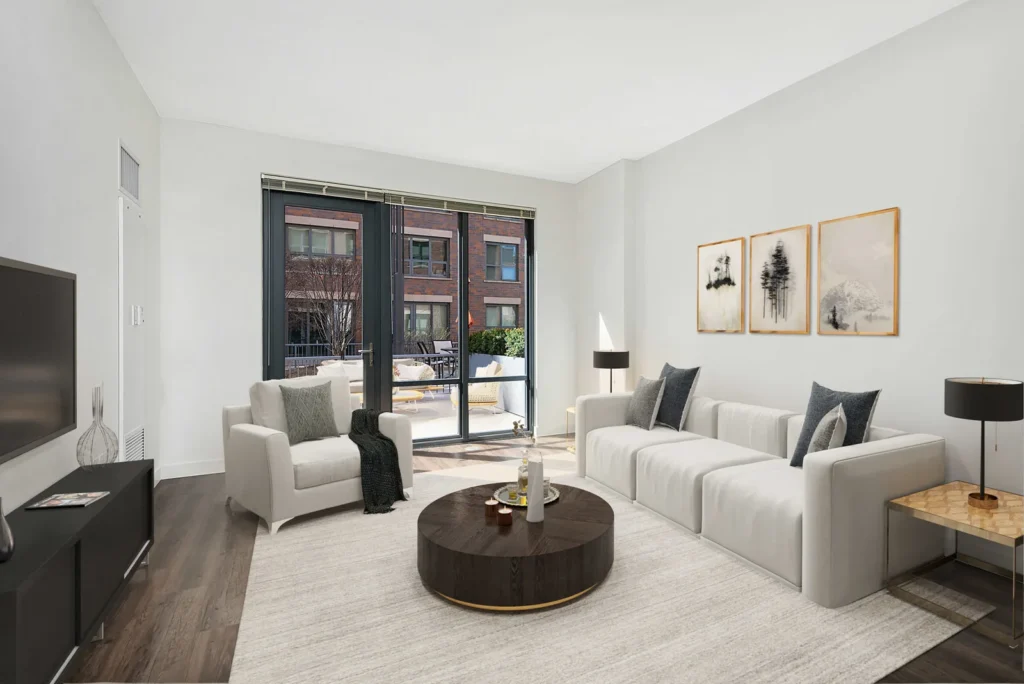 Modern living room with white walls, two white sofas, a round dark coffee table, wall art, TV, and large glass doors leading to a balcony with an outdoor seating area.