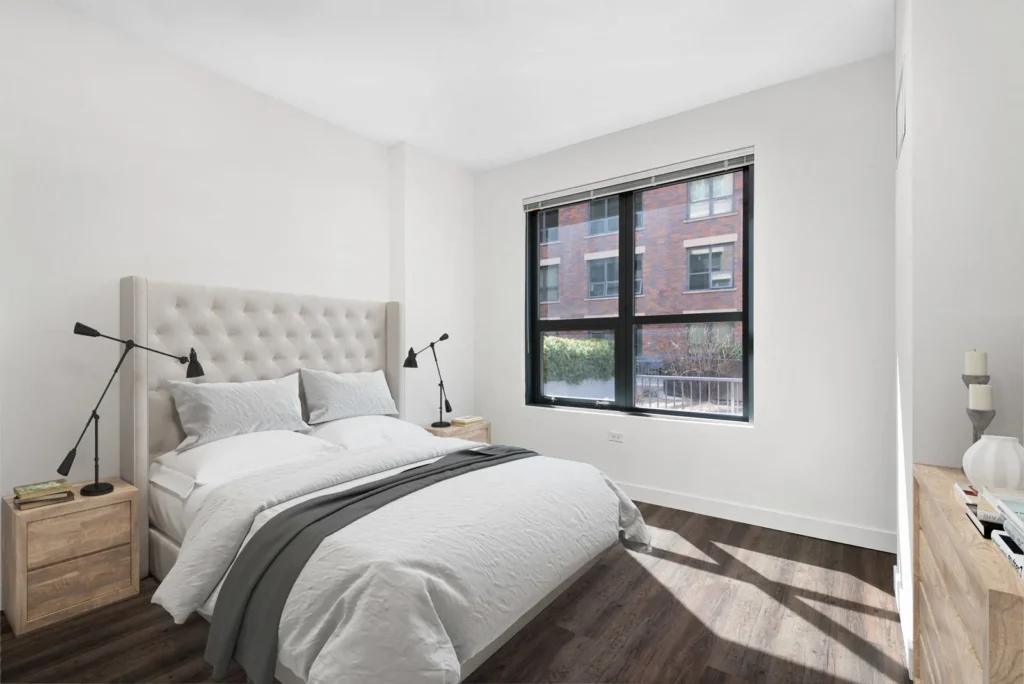 A modern bedroom with a large bed, two bedside tables with lamps, a dresser, white walls, dark wood flooring, and a window overlooking a brick building.