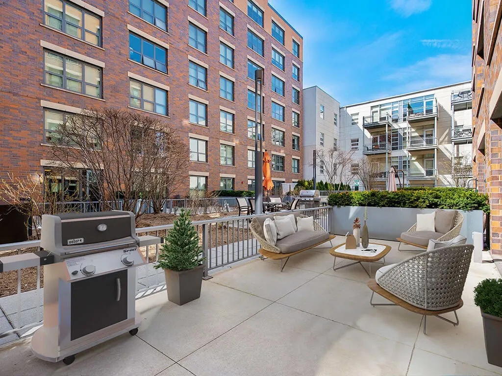 Patio area with outdoor seating, table, potted plant, and grill, surrounded by apartment buildings under a clear blue sky.