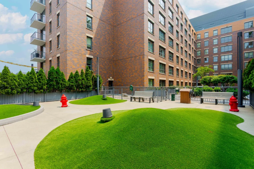 A modern apartment complex courtyard features artificial grassy mounds, red fire hydrants, a bench, trees along a fence, and multi-story brick buildings in the background.