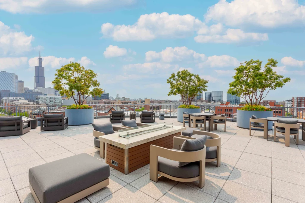 Rooftop patio with modern outdoor seating, a fire pit table, large potted trees, and a city skyline view under a partly cloudy sky.