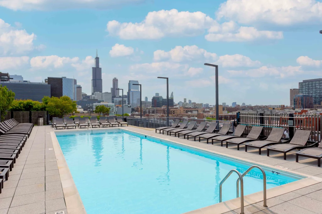 Rooftop outdoor swimming pool with lounge chairs, city skyline and high-rise buildings visible in the background under a partly cloudy sky.