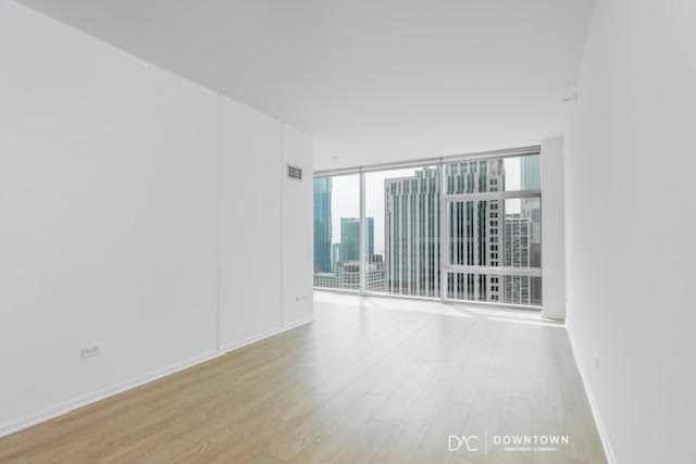 Unfurnished high-rise apartment with light wood flooring, white walls, and floor-to-ceiling windows overlooking a city skyline with modern office buildings.