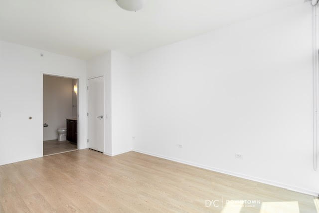 Empty, unfurnished room with white walls, light wood flooring, a door leading to a bathroom, and a closed white door on the left wall.