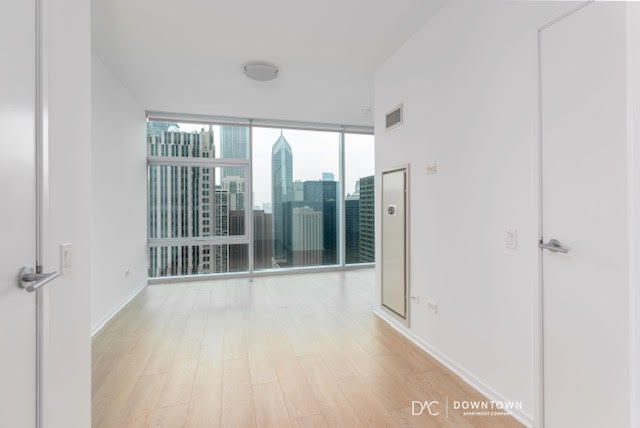 Bright empty room with light wood flooring, white walls, and floor-to-ceiling windows offering a city skyline view with tall buildings.