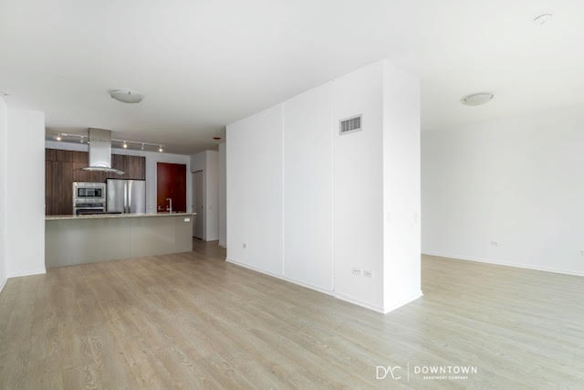 Spacious, unfurnished modern apartment with light wood flooring, an open kitchen featuring stainless steel appliances, and white walls throughout.