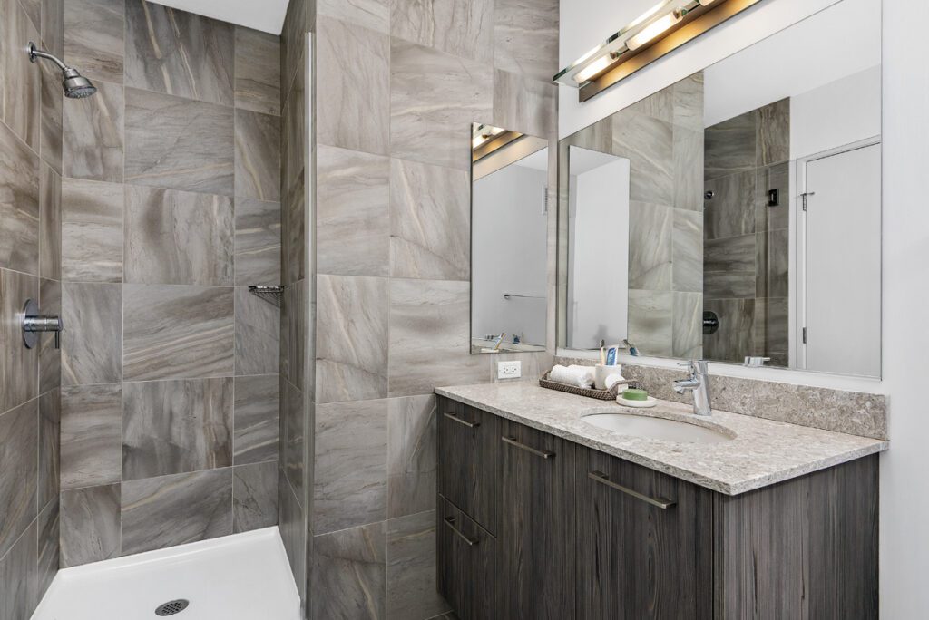 Modern bathroom with a tiled walk-in shower, large mirror, marble countertop, and wooden vanity with drawers.