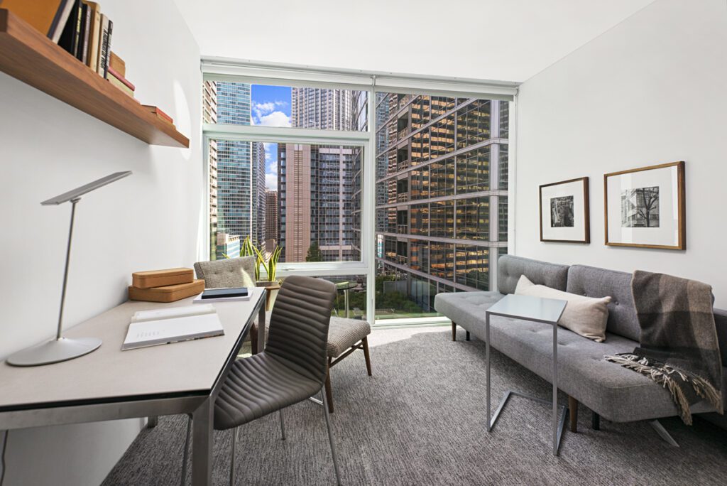 A modern, compact office and living space with a desk, chair, sofa, wall art, and large windows overlooking tall city buildings.
