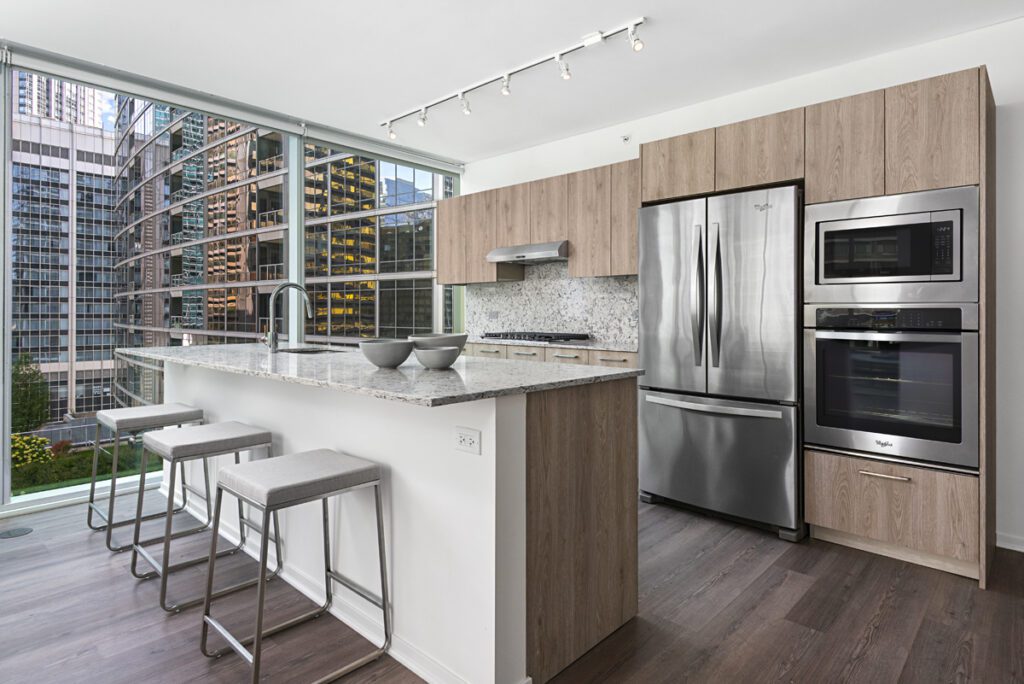 Modern kitchen with light wood cabinets, stainless steel appliances, and a marble island with three barstools; large windows show city buildings outside.
