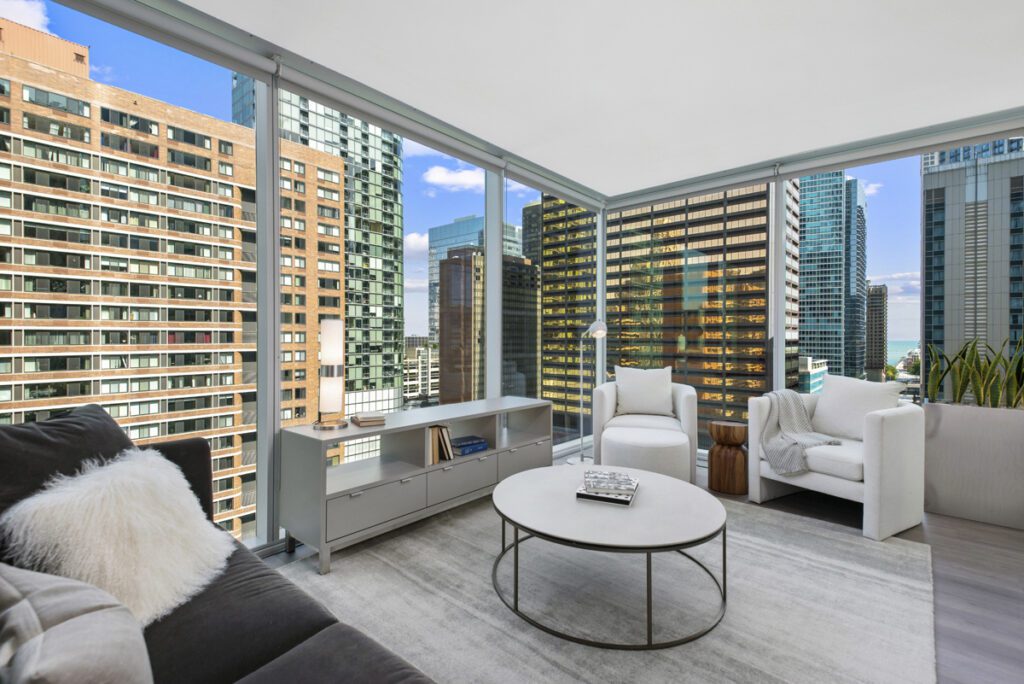 Modern living room with large windows showcasing a city skyline, featuring neutral-toned furniture, a round coffee table, and a minimalist shelving unit.
