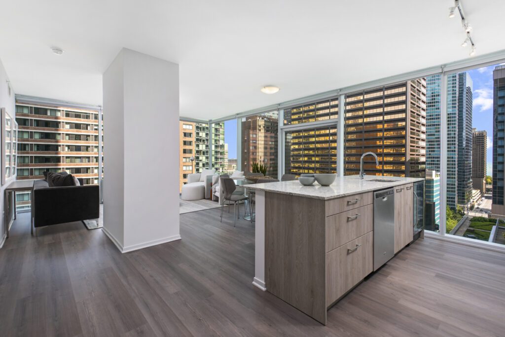 Modern open-concept apartment with large windows, kitchen island, living and dining area, and views of city skyscrapers.