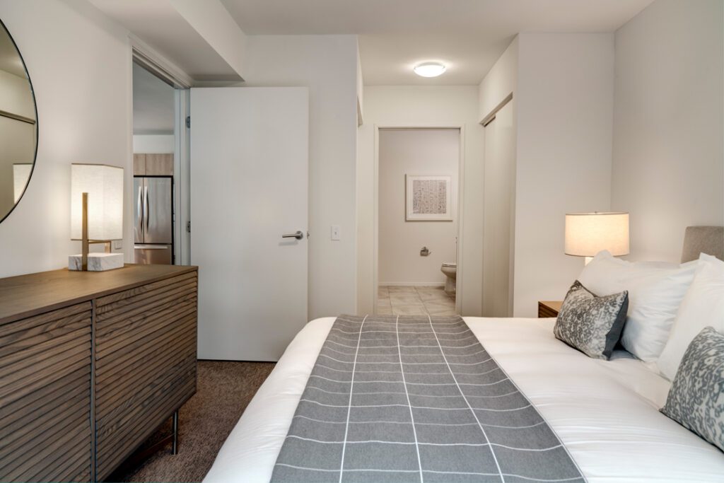 Modern bedroom with a double bed, two lamps, a dresser with a round mirror, and an open door leading to a bathroom with visible toilet and wall art.
