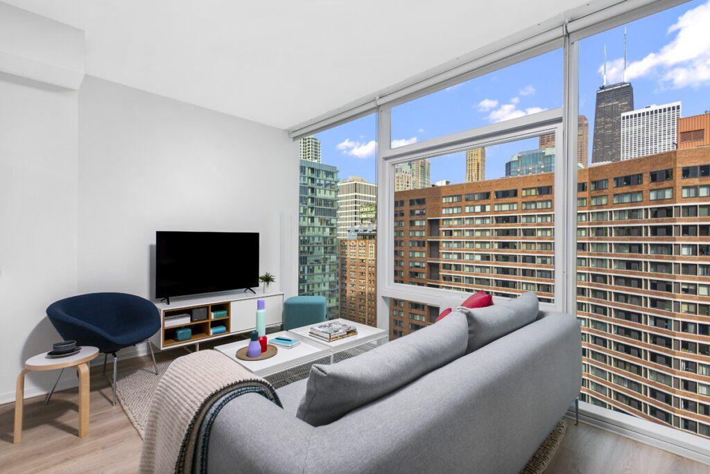 Modern living room with a gray sofa, TV, small table, and large windows showing a city skyline with tall buildings and blue sky.