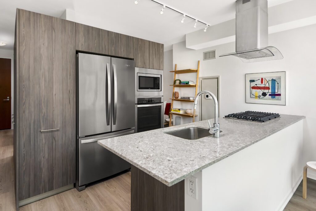 Modern kitchen with stainless steel appliances, gray wood cabinets, a granite island with sink, gas cooktop, and open shelving against a white wall with abstract art.