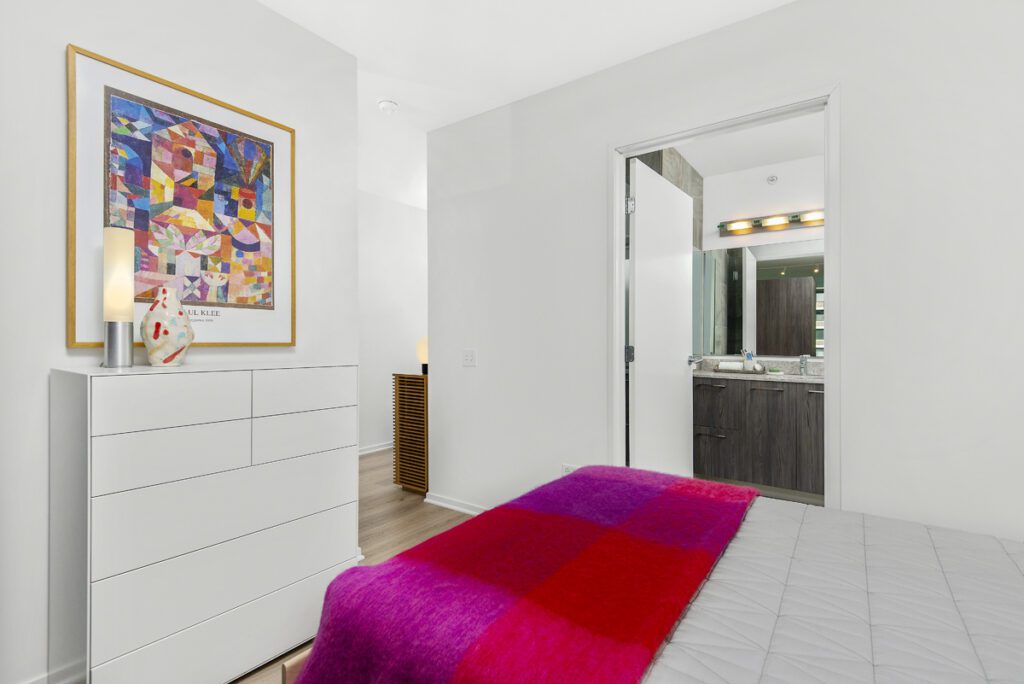 A bedroom features a bed with a red and purple blanket, a white dresser with art above it, and an open door leading to a modern bathroom.