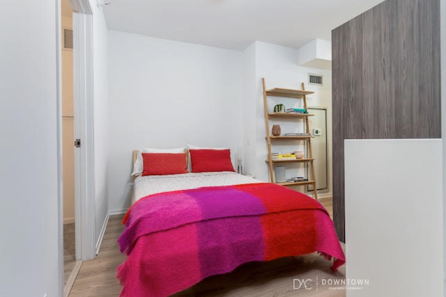 A small bedroom with a double bed, red and pink blanket, two red pillows, a wooden shelf with decor, and white walls.