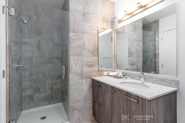 Modern bathroom with a glass-enclosed walk-in shower, large mirror, granite countertop, and wooden vanity with drawers. Bright lighting above the mirror.