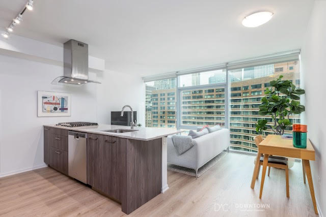Modern apartment interior with an open kitchen, living area, large windows, city view, and a small desk with a plant.