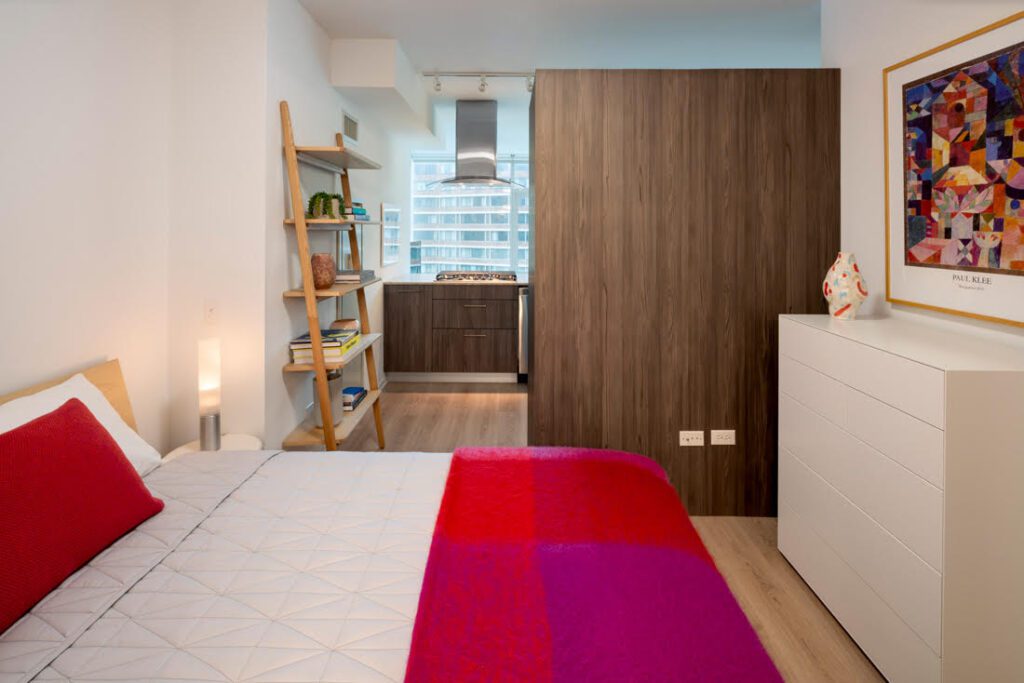 Modern studio apartment with a bed, colorful bedding, wooden shelving, dresser, framed artwork, and view of a kitchen separated by a wood partition.