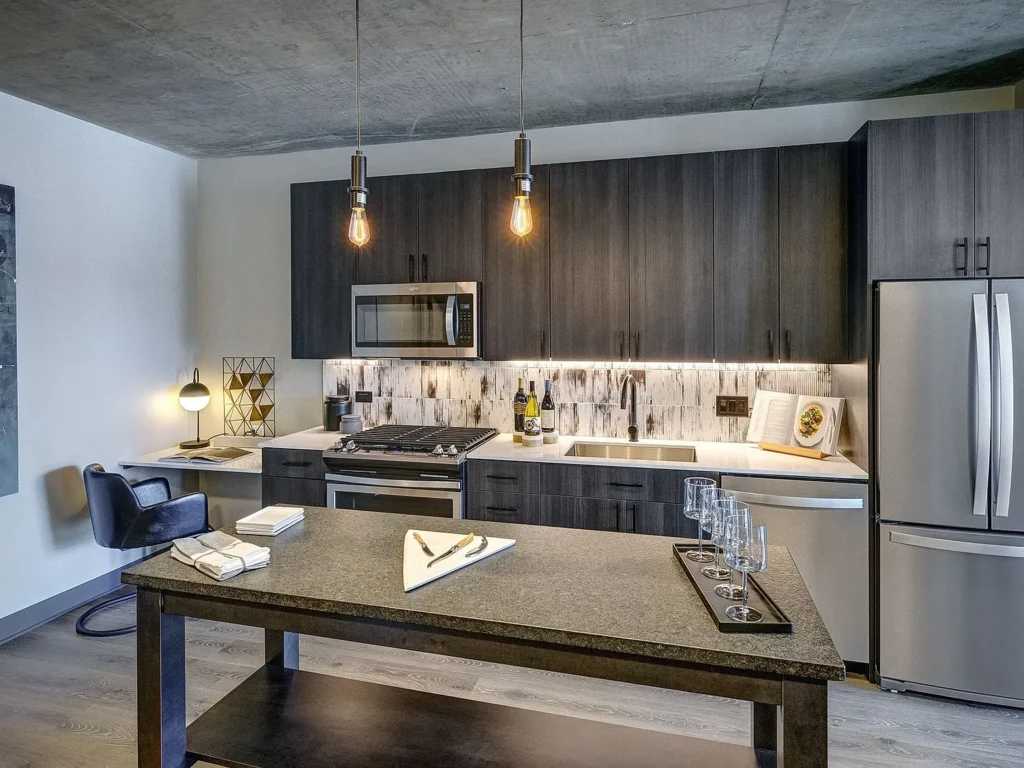 Modern kitchen with dark wood cabinets, stainless steel appliances, marble backsplash, island with barware, and two pendant lights hanging from a concrete ceiling.