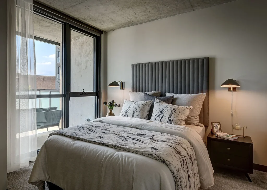 Modern bedroom with a large bed, gray upholstered headboard, bedside tables with lamps, and floor-to-ceiling windows leading to a balcony.