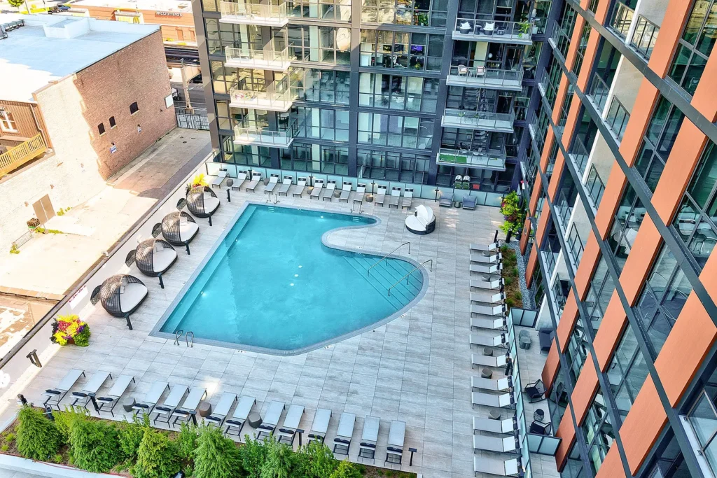Aerial view of a modern apartment complex pool area with lounge chairs, sun umbrellas, and adjacent high-rise buildings.