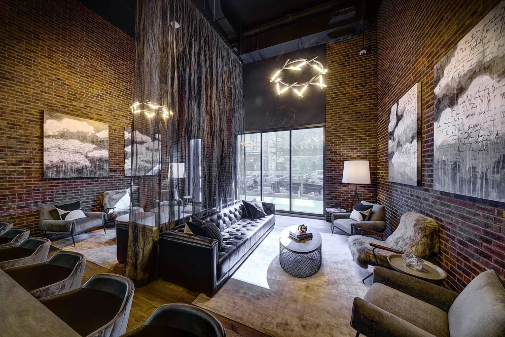 Modern lounge with exposed brick walls, contemporary furniture, abstract art, hanging light fixtures, and a sheer curtain divider near large windows.