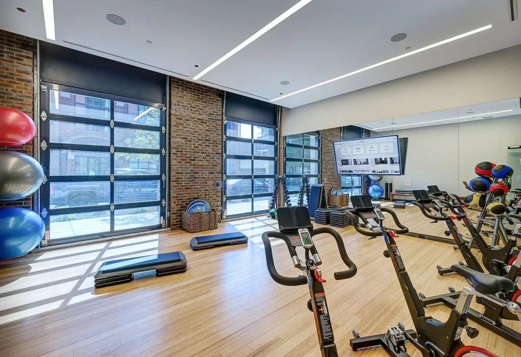 Modern gym with stationary bikes, exercise mats, stacked step platforms, stability balls, and large windows with brick walls allowing natural light in.