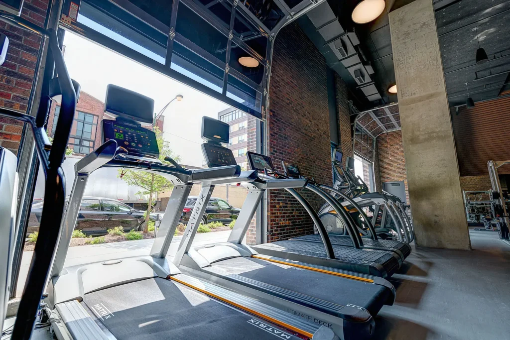 Row of treadmills facing large open garage-style windows in a modern gym with exposed brick and concrete walls, allowing natural light to enter.