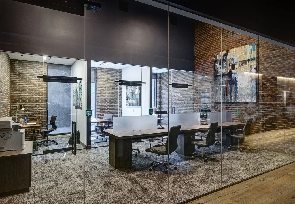 Modern office with brick walls, glass partitions, several desks and chairs, computers, and abstract art on the wall, viewed from outside the glass enclosure.