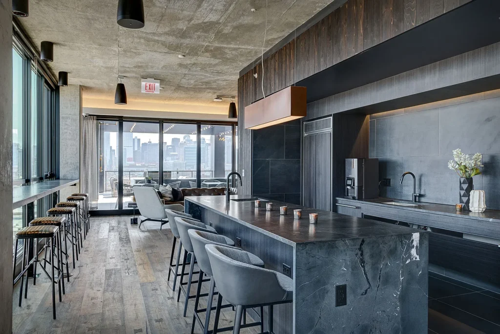 Modern kitchen and lounge area with dark marble island, gray seating, bar stools by large windows, and city skyline visible through glass doors to a balcony.