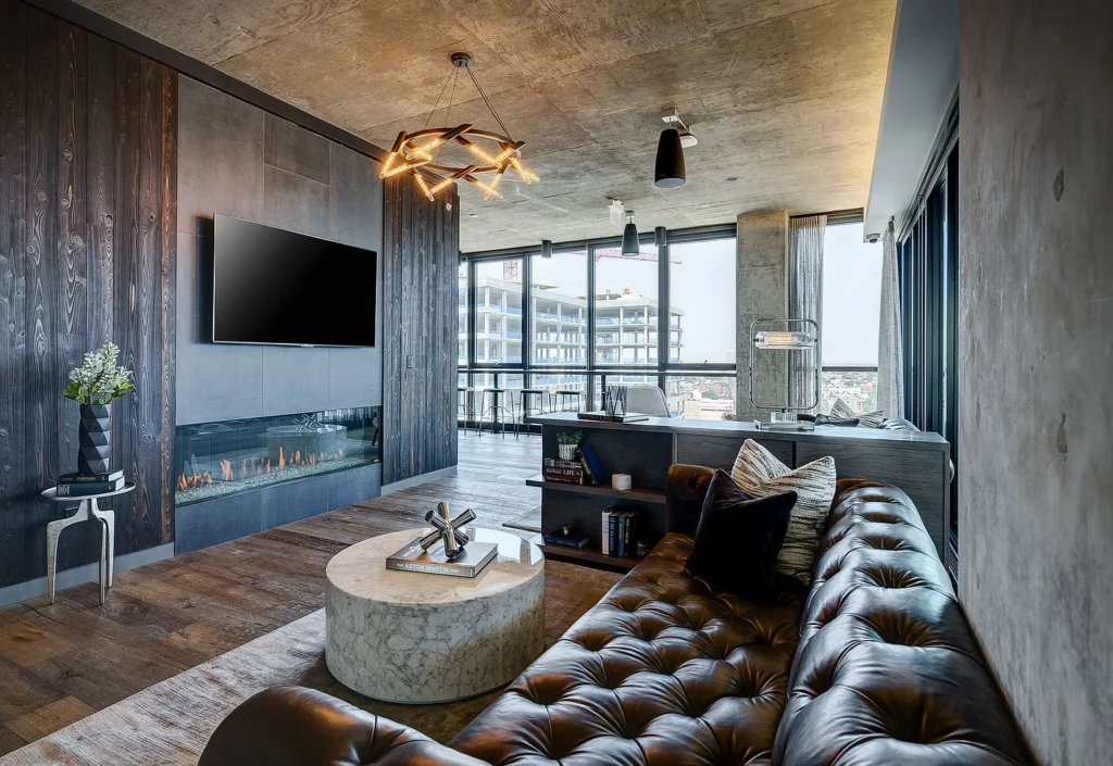 Modern living room with a leather sofa, marble coffee table, wall-mounted TV, and electric fireplace; large windows reveal a city view and construction outside.
