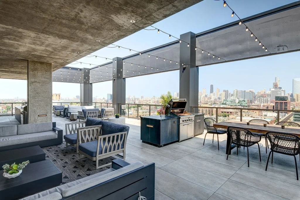 Rooftop patio with lounge seating, dining table, grill station, and string lights, offering city skyline views under a clear sky.