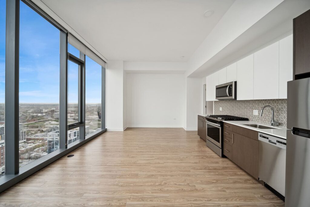Modern apartment kitchen with stainless steel appliances, wood flooring, and floor-to-ceiling windows offering a city view.