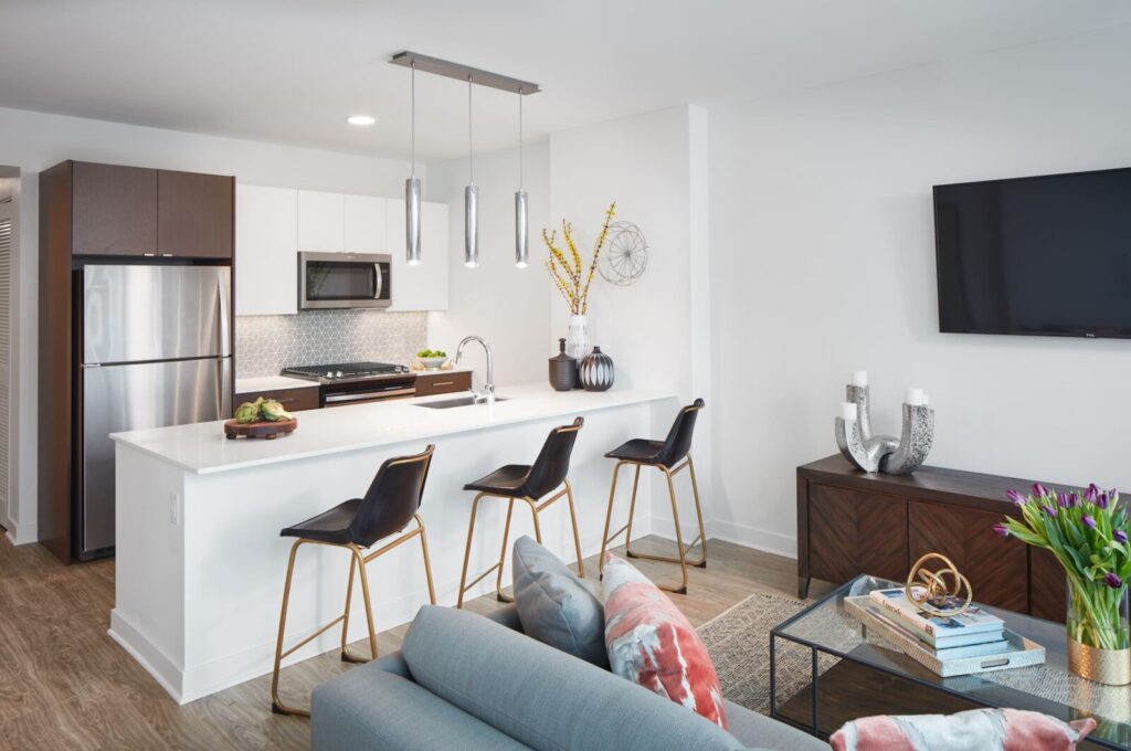 Modern apartment kitchen and living area with a white island, three black bar stools, stainless steel appliances, and a wall-mounted TV.