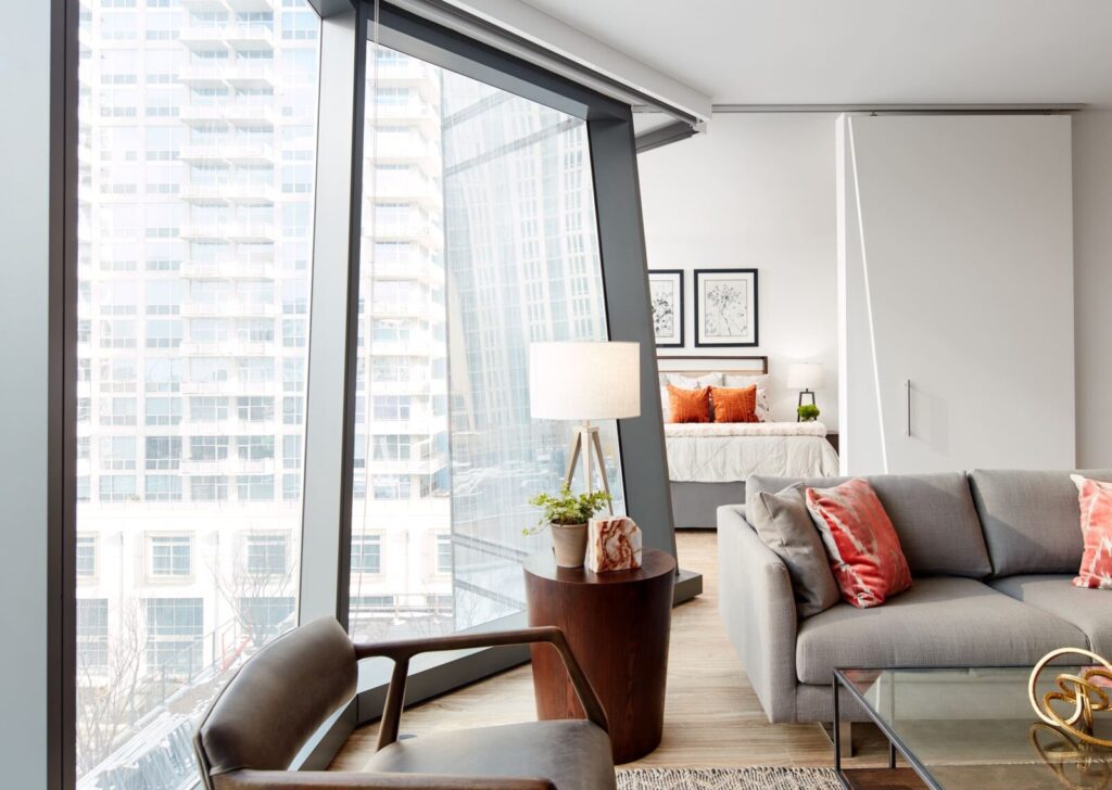 Modern apartment with large angled windows, a grey sofa with orange pillows, a glass coffee table, and a view into a bedroom with a neatly made bed and framed art on the wall.