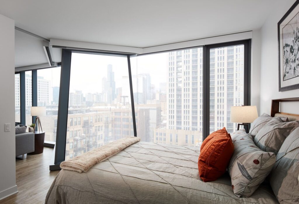 Modern bedroom with large slanted windows offering a city skyline view, featuring a neatly made bed with patterned pillows and a lamp on a bedside table.